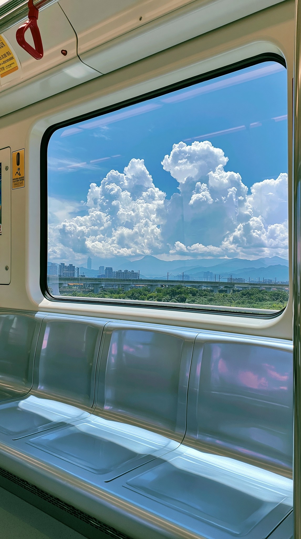 晴天夏日的轻轨车厢内景 空无一人的城市轨道交通车厢 正对大面积车窗拍摄 窗外是通透蓝天 蓬松白云 远处城市高架 绿色植被和淡蓝色远山 车厢内有银灰色金属长椅 座椅表面反射蓝天光影...